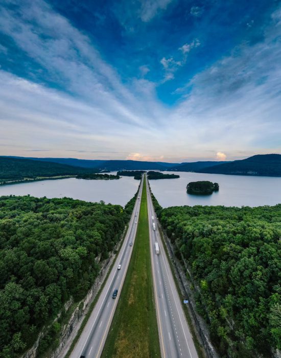 A breathtaking aerial shot of a highway running through lush forests beside a serene lake at sunset.